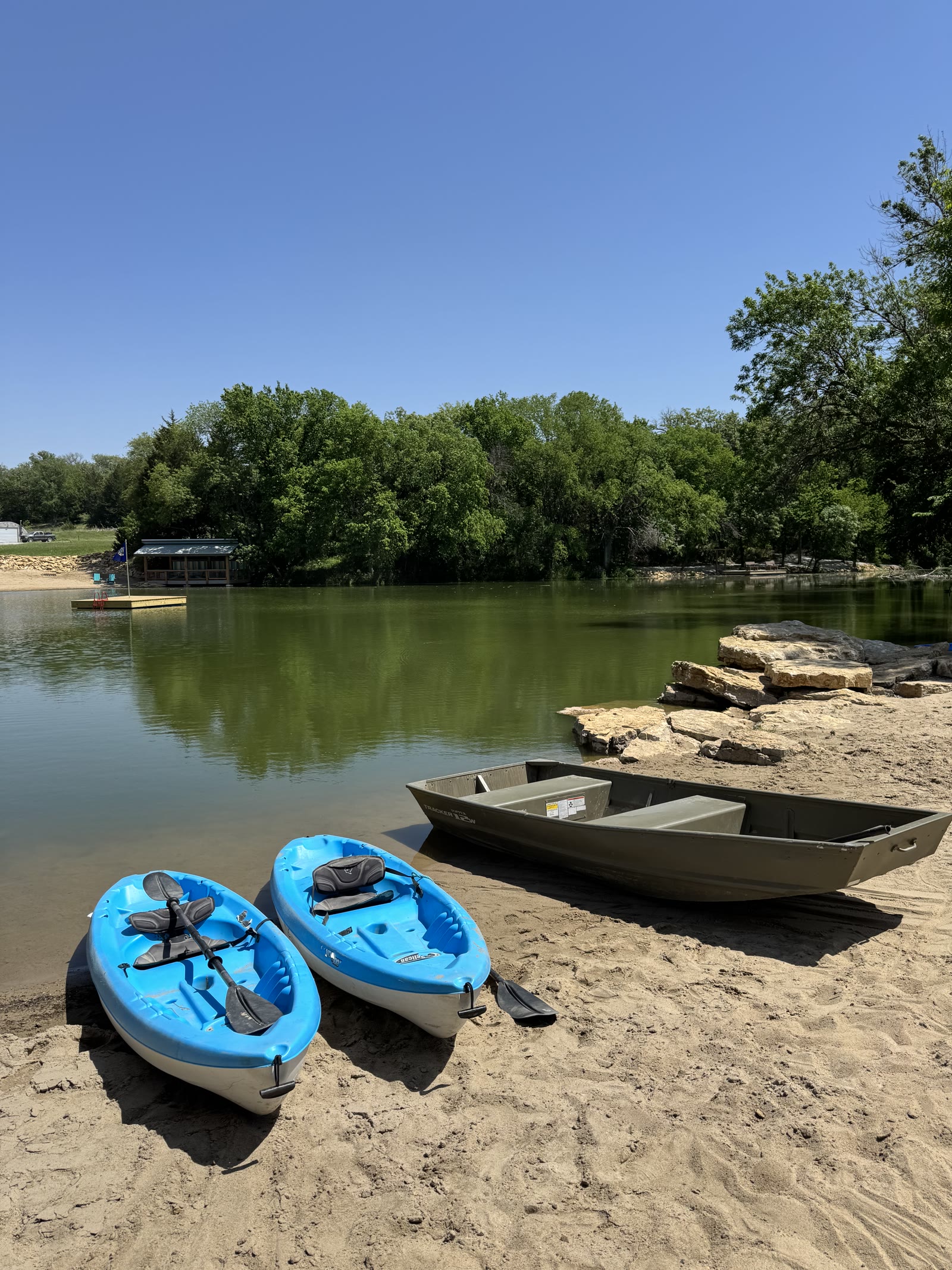Kayaks and a jon boat on the sandy beach of the stocked lake, swim platform beyond